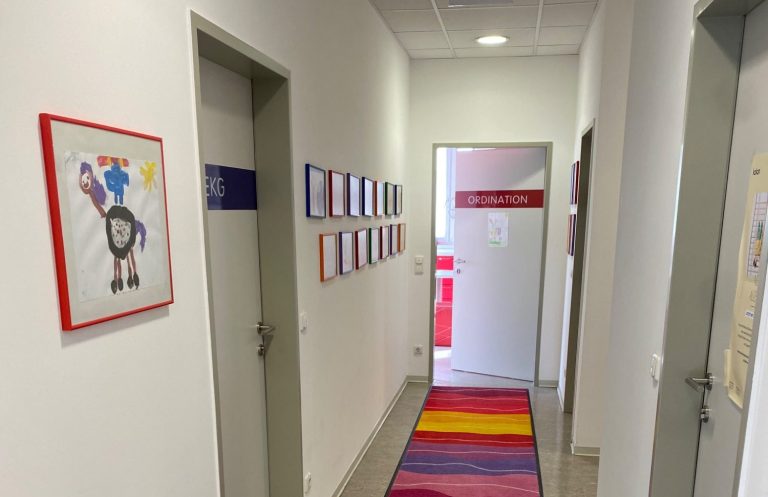 Hier geht es zum Kinderkardiologen Dr. Prandstetter Blick in einen hellen Flur mit bunten Zeichnungen an der Wand und einem bunten Teppich.