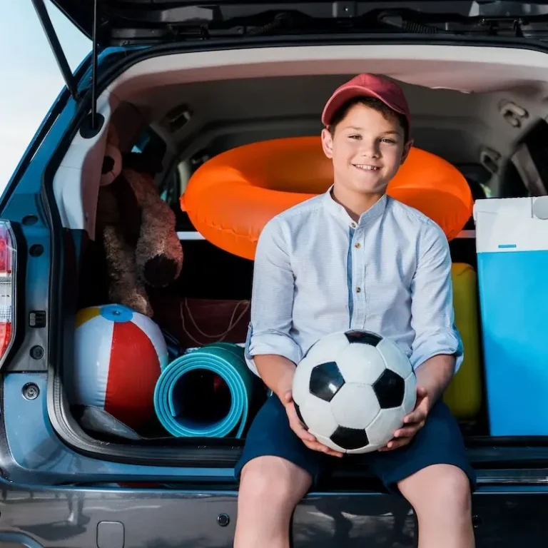 Ab in die Kinderherzpraxis - mit dem Auto Ein Junge sitzt im Kofferraum eines Autos und hält einen Fußball, umgeben von Spielzeugen.