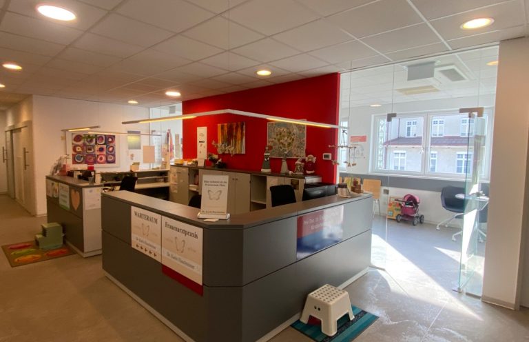 Willkommen in der Kinderherzpraxis Linz Empfang der Kinderherzpraxis Linz mit roter Wand und Empfangstresen in einer hellen Umgebung.