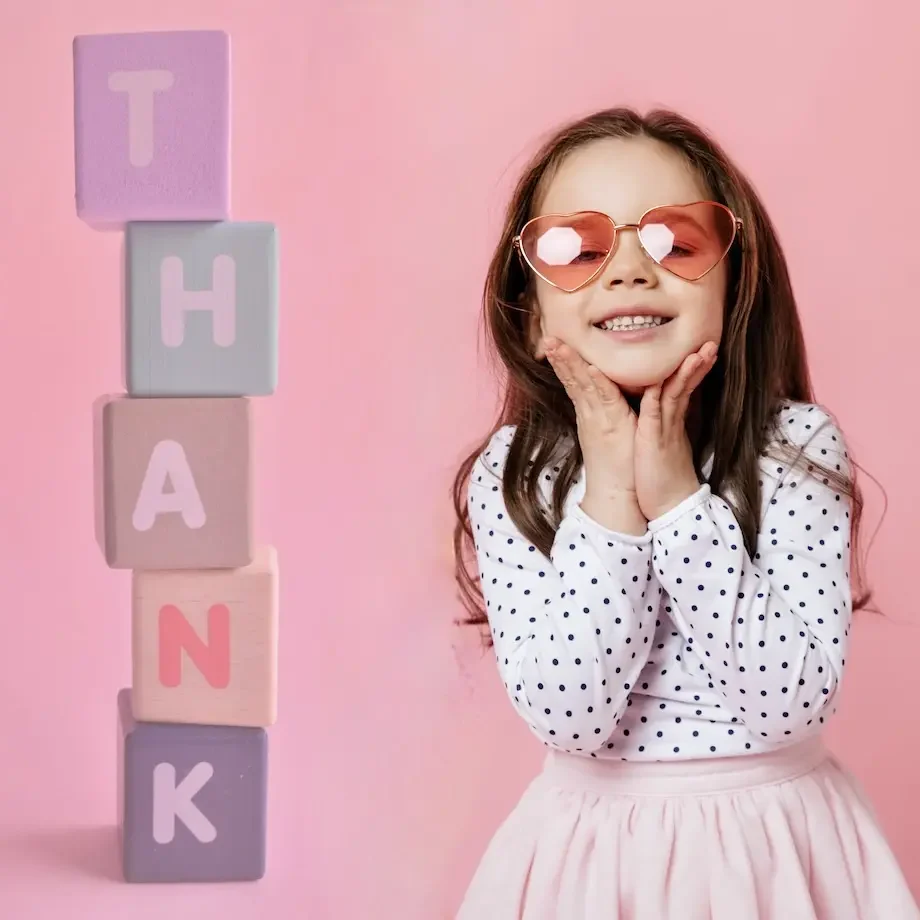 Ein Mädchen mit Sonnenbrille lächelt, umgeben von bunten Bauklötzen mit dem Wort „THANK“.